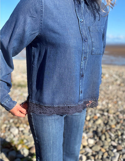 A person outdoors wearing a dark blue denim button-up shirt with a lace hem detail, paired with blue jeans, standing on a rocky beach.