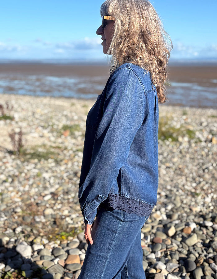 A woman standing on a rocky beach wearing a blue denim jacket with a dark lace hem detail and matching blue jeans, photographed from the side profile against a blurred background of water and sky.