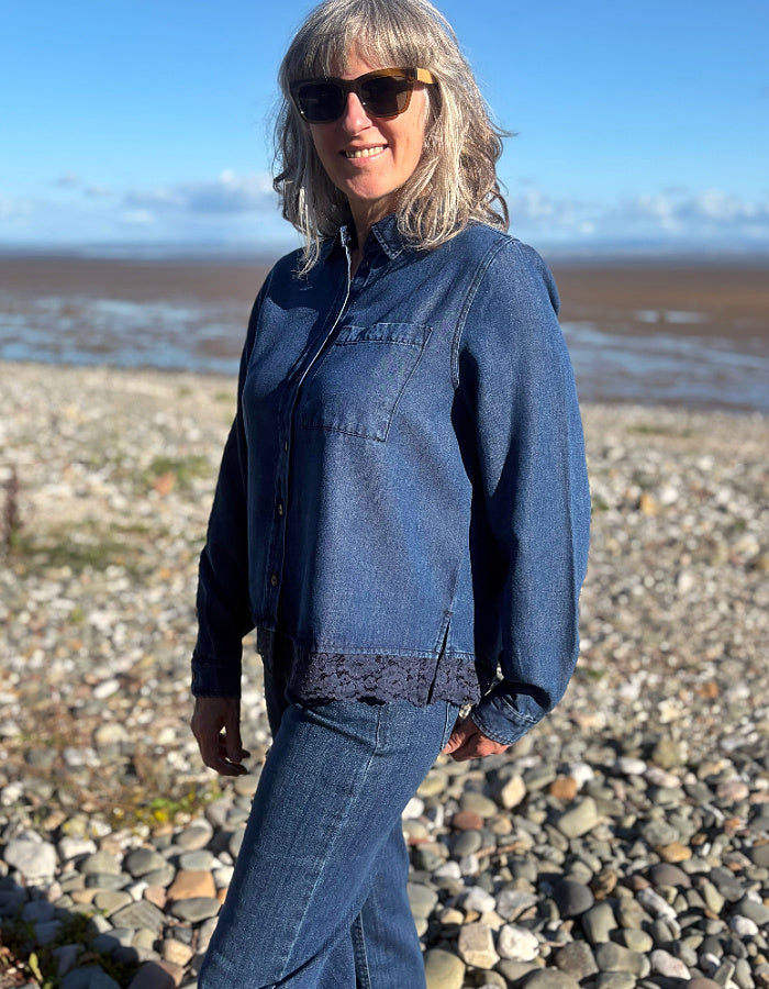 A woman wearing a blue denim button-up shirt and matching denim pants stands on a rocky beach with water and sky in the background.