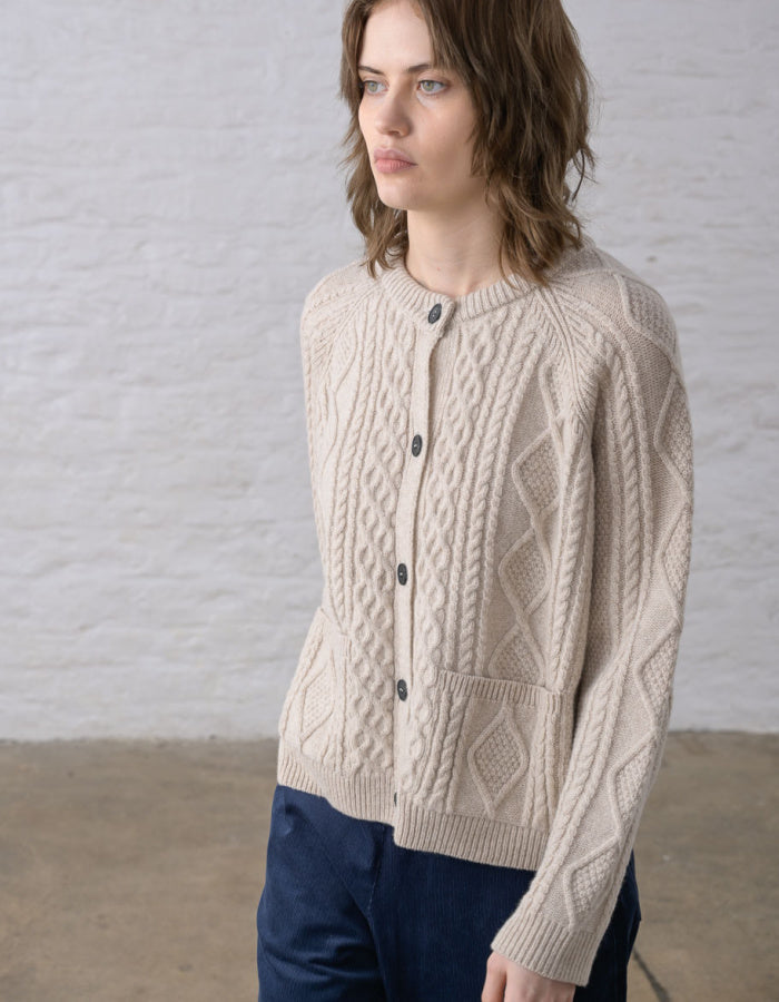 Close-up of a person wearing a cream cable-knit Aran cardigan sweater with dark buttons, against a soft light grey background.
