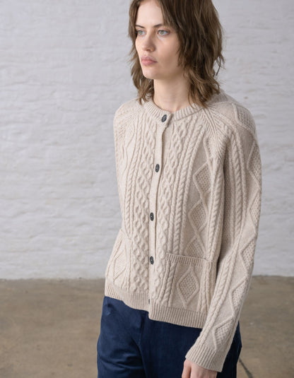 Close-up of a person wearing a cream cable-knit Aran cardigan sweater with dark buttons, against a soft light grey background.
