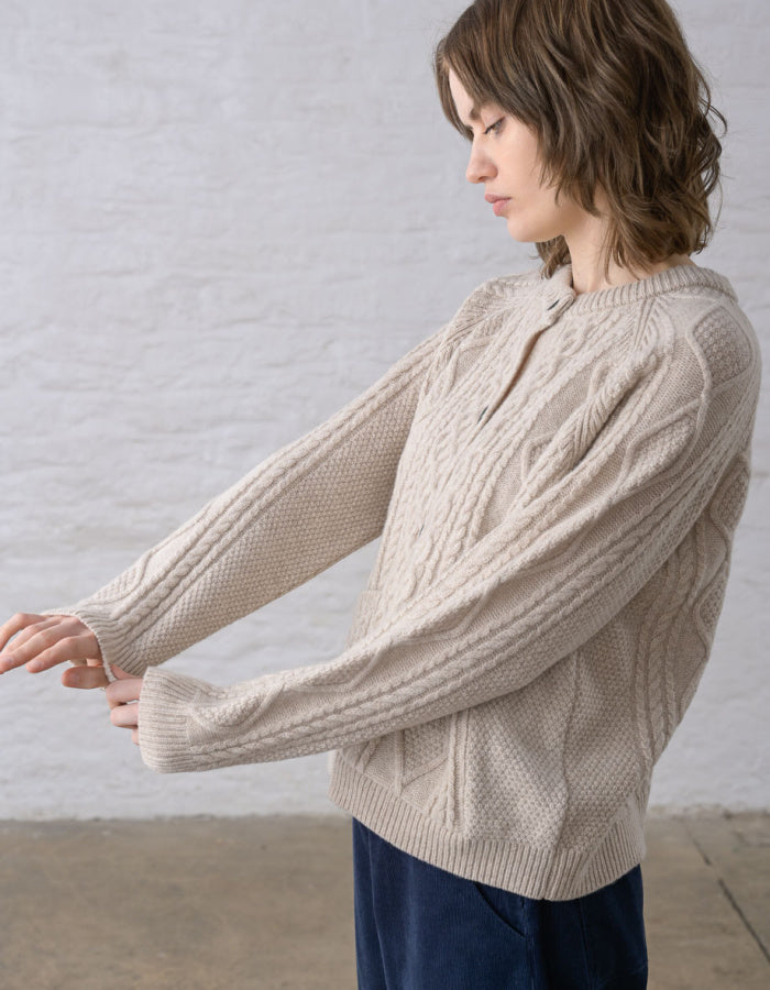 Close-up of a person wearing a cream cable-knit Aran cardigan sweater with dark buttons, against a soft light grey background.
