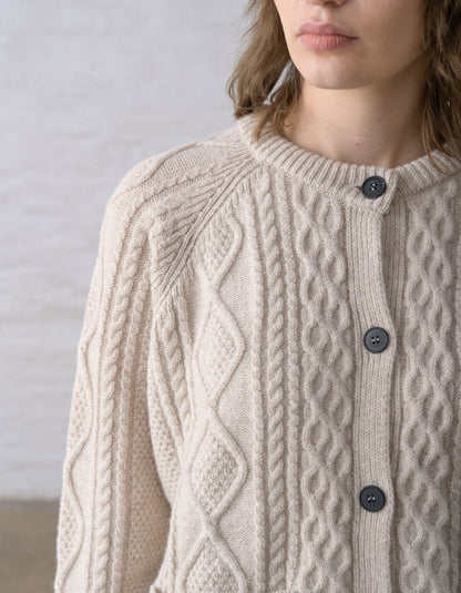 Close-up of a person wearing a cream cable-knit Aran cardigan sweater with dark buttons, against a soft light grey background.