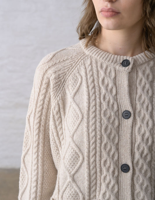 Close-up of a person wearing a cream cable-knit Aran cardigan sweater with dark buttons, against a soft light grey background.