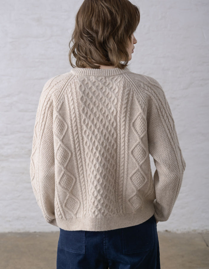 A person shown from the back wearing a cream cable-knit sweater and dark blue jeans, standing against a white textured wall.