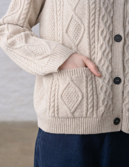 Close-up of a person wearing a cream cable-knit Aran cardigan sweater with dark buttons, against a soft light grey background.