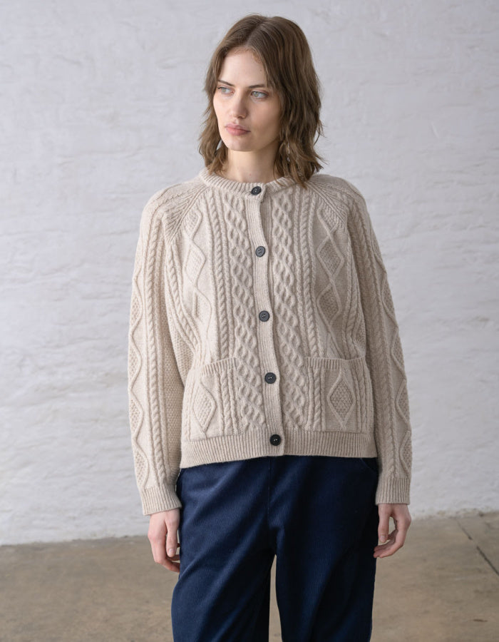 A woman wearing a beige cable-knit cardigan sweater with dark buttons and navy blue trousers, standing against a white textured wall.