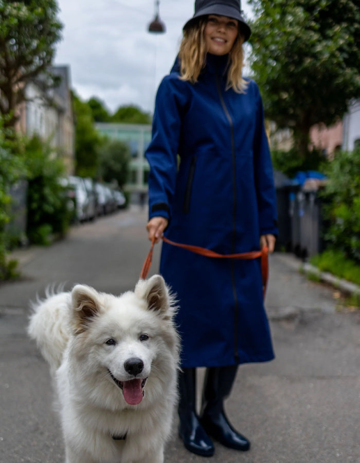 A woman wearing a long navy blue waterproof coat, black hat, and black rubber boots walks a fluffy white Samoyed dog on a street.