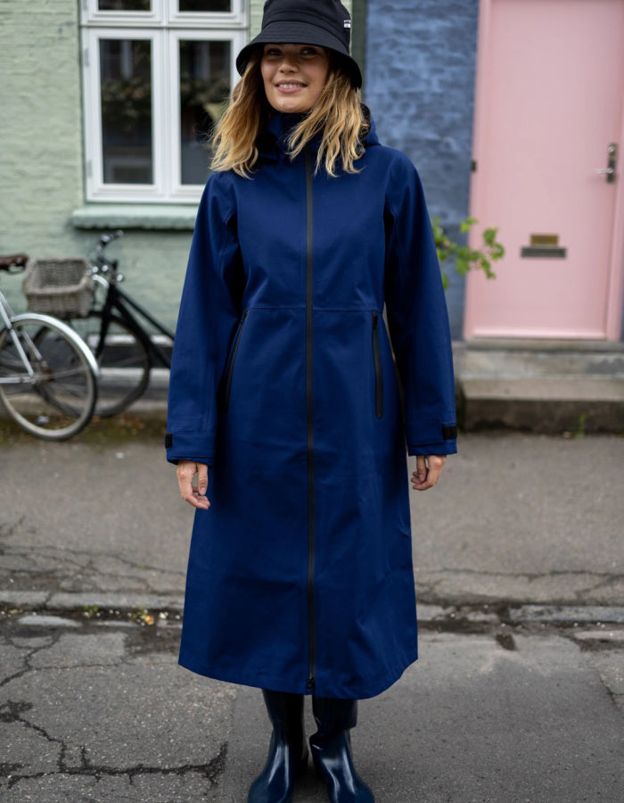 A woman wearing a long navy blue raincoat, a black bucket hat, and dark rain boots stands outdoors on a street in front of a building with a pink door and a bicycle.