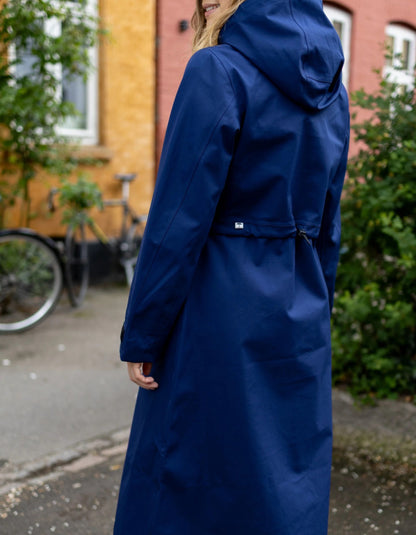A woman wearing a long navy blue hooded raincoat, photographed from the back in an outdoor urban setting with a brick building and bicycle in the background.