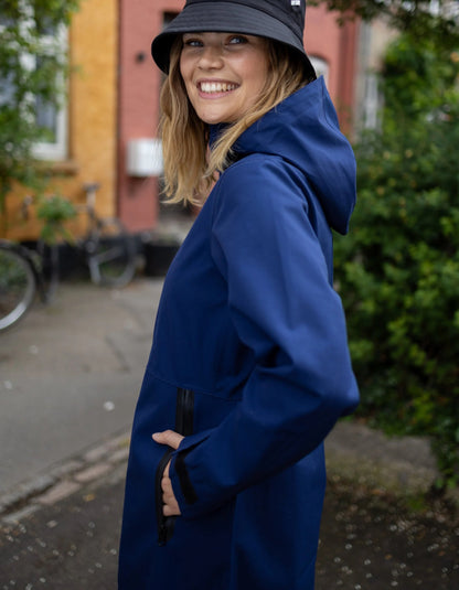 A woman wearing a navy blue hooded raincoat and a black bucket hat, smiling while looking back over her shoulder in an outdoor urban setting with a sidewalk, bicycle, and brick building in the background.