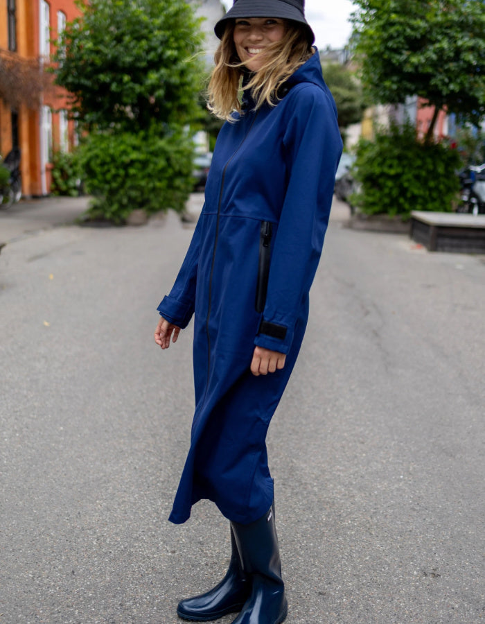 A woman wearing a long navy blue hooded raincoat and matching navy rain boots, standing on an outdoor street with trees and buildings in the background.