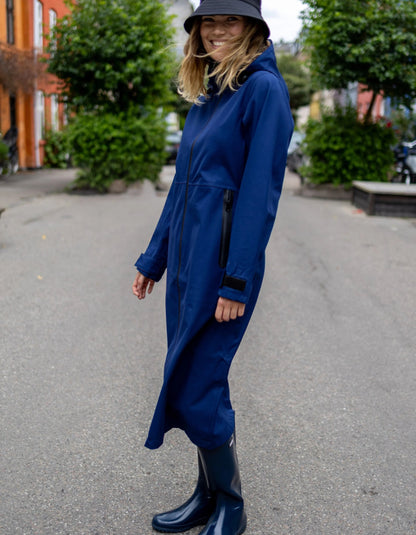 A woman wearing a long navy blue hooded raincoat and matching navy rain boots, standing on an outdoor street with trees and buildings in the background.