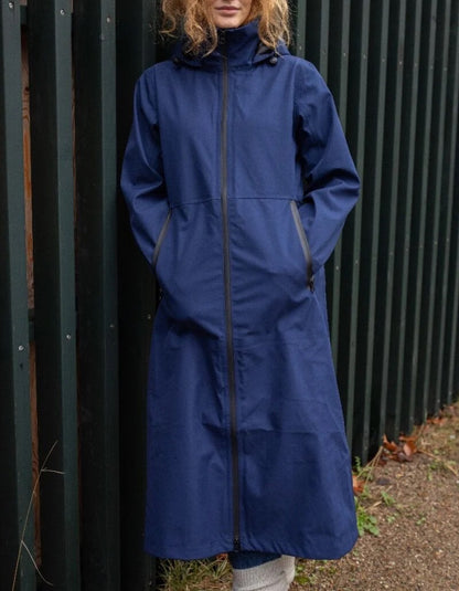 A woman wearing a long navy blue hooded raincoat with a full front zipper, standing in front of a dark green corrugated metal fence.