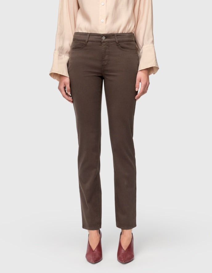 A model wearing a pair of straight-leg brown pants, paired with a light beige blouse and burgundy pointed-toe heels, shown from the waist down on a light gray background.