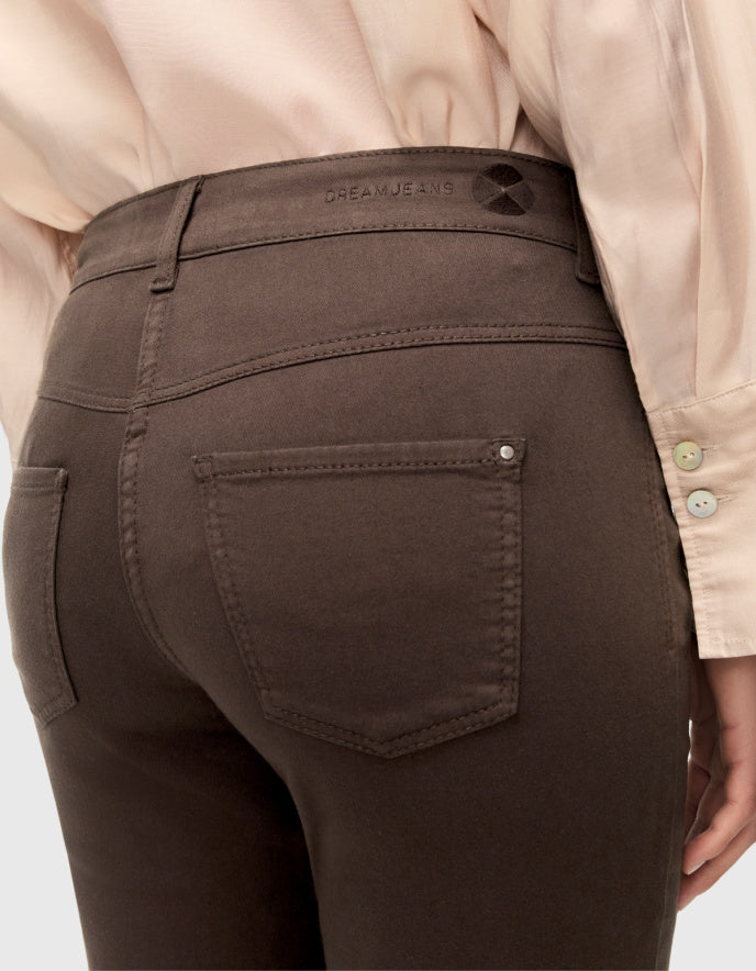 Close-up back view of a person wearing dark brown slim-fit jeans with a light beige button-up shirt, showing the back pocket and waistband with 'DREAM JEANS' branding.