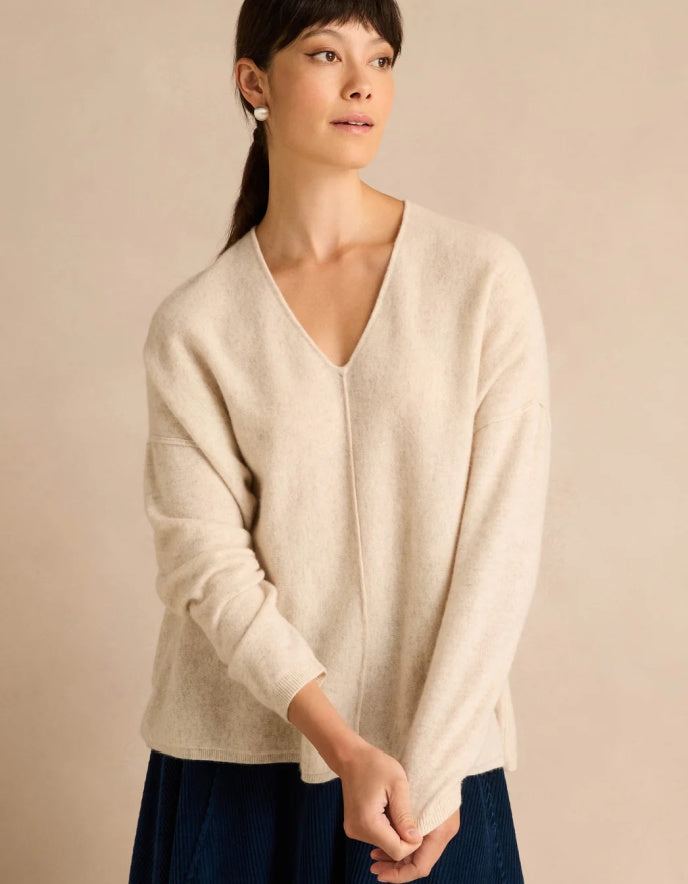 A woman wearing a cream-colored, V-neck knit sweater and a dark blue skirt, standing against a solid beige background.