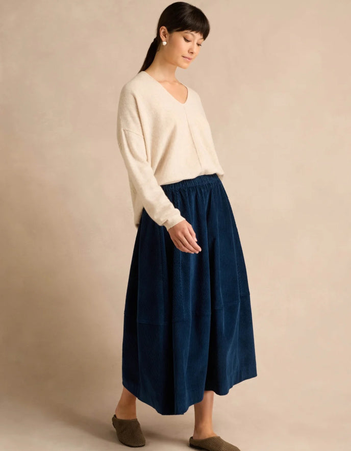 A woman wearing a cream-colored knit sweater and a dark navy blue midi skirt, standing against a solid beige studio background.