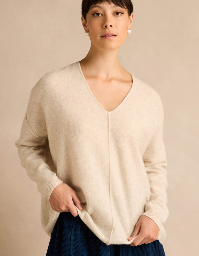 A woman wearing a cream-colored V-neck knit sweater, paired with a dark blue corduroy skirt, standing against a solid beige background.
