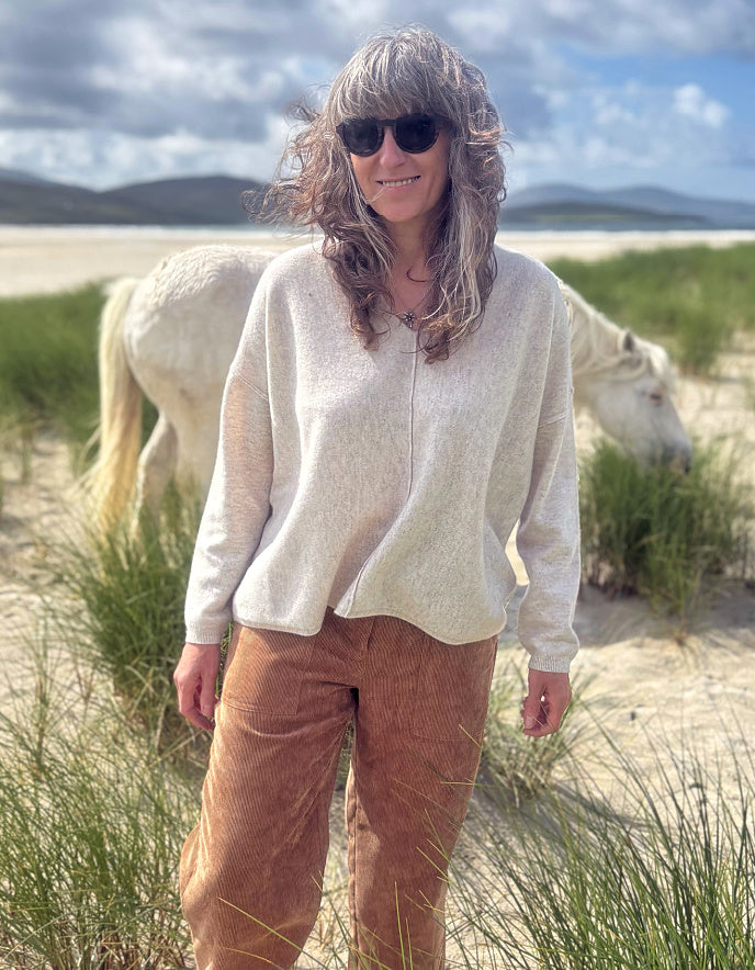 A woman wearing a cream-colored knit sweater and brown corduroy pants stands in a grassy beach dune setting with a white horse in the background.