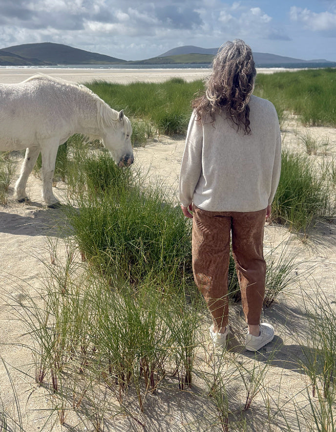 A woman stands on a sandy beach dune wearing a cream knit sweater and brown corduroy pants, looking out towards a lake with mountains in the background. A white horse grazes nearby among green beach grass.
