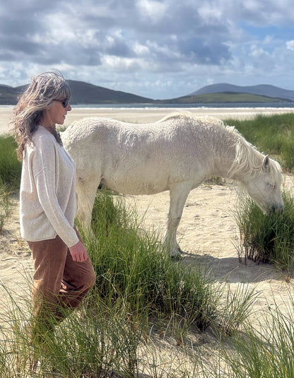 A woman wearing a cream knit sweater and brown pants stands on a sandy beach next to a white horse grazing in green dune grass, with mountains and a cloudy sky in the background.