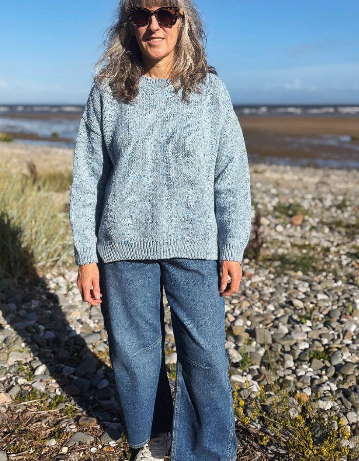 A woman wearing a light blue donegal tweed merino knit sweater and blue jeans stands on a rocky beach with water and sky in the background.