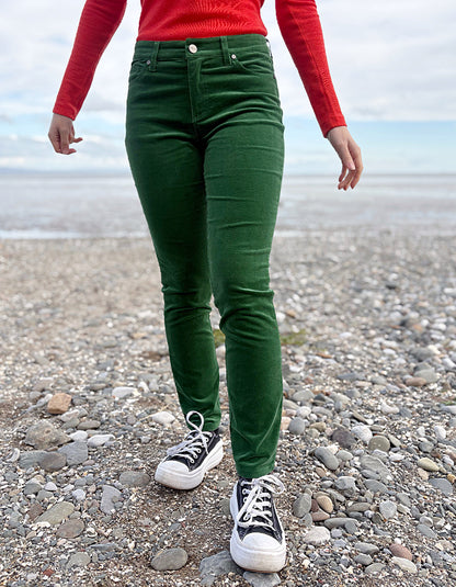 Person wearing green velvet pants and red sweater standing on a pebbly beach.