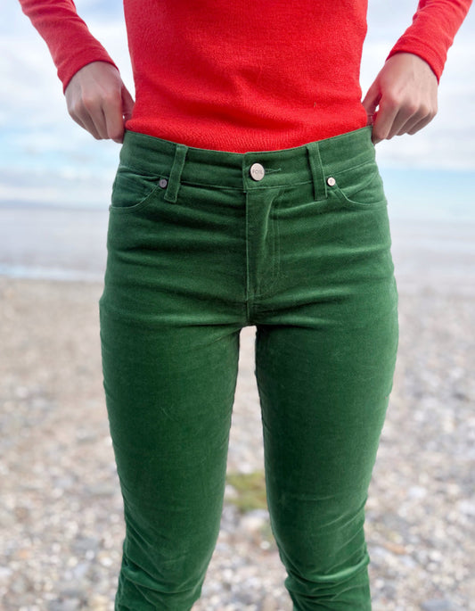 Person wearing green velvet pants and red sweater standing on a pebbly beach.