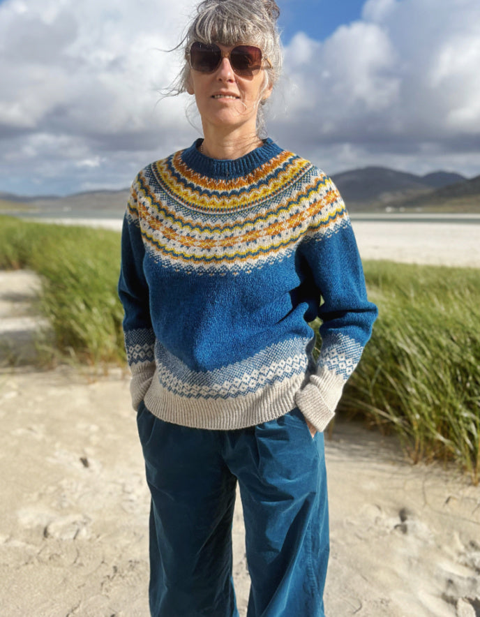 A woman wearing a blue Fair Isle knit sweater with yellow, orange, and white patterns, standing outdoors on a sandy path with grass and mountains in the background.