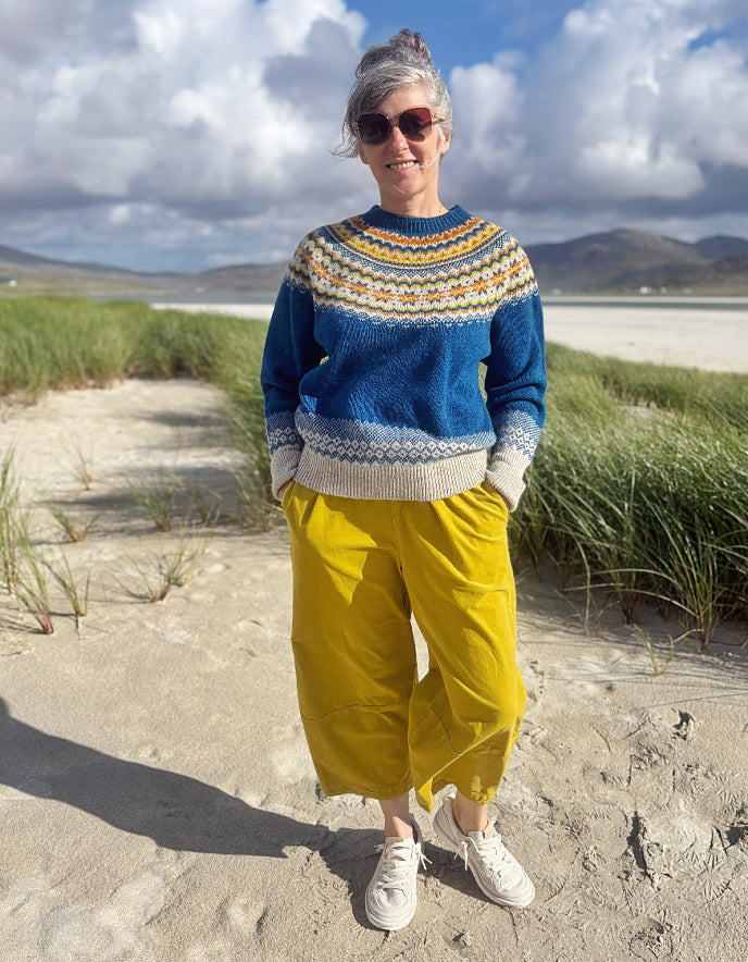 A woman standing on a sandy path outdoors, wearing a blue and cream Fair Isle knit sweater, bright yellow cropped pants, white sneakers, and sunglasses, with grassy dunes and a cloudy sky in the background.