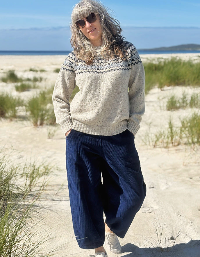 A woman standing on a sandy beach wearing a cream knit sweater with a dark navy patterned yoke and dark navy wide-leg pants.