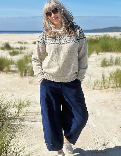 A woman standing on a sandy beach wearing a cream knit sweater with a dark navy patterned yoke and dark navy wide-leg pants.