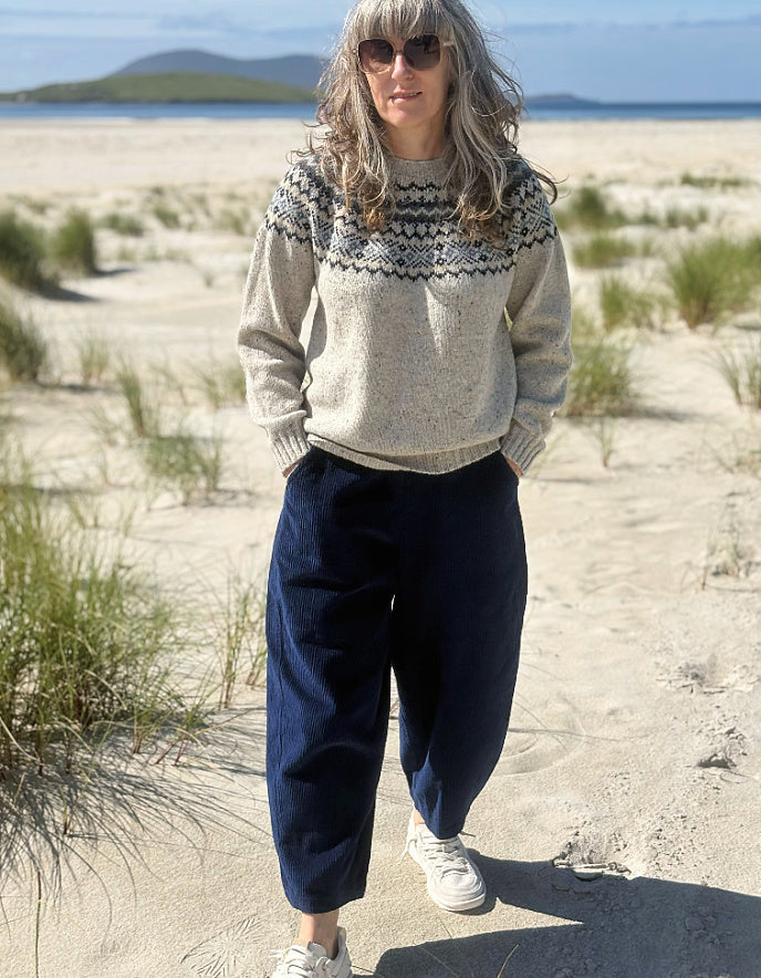 A woman standing on a sandy beach wearing a light beige knit sweater with a dark geometric pattern across the chest, navy blue wide-leg pants, and white sneakers.