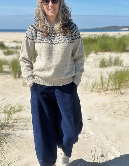 A woman standing on a sandy beach wearing a cream knit sweater with a dark geometric yoke pattern and dark navy wide-leg pants.