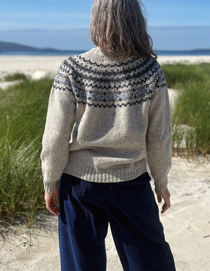 A person standing on a sandy beach facing away from the camera, wearing a cream-colored knit sweater with a dark gray and black geometric Fair Isle yoke pattern, paired with dark blue corduroy pants.