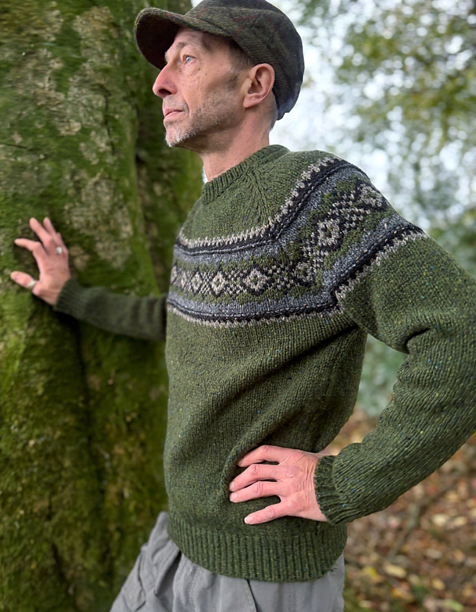 Man wearing a green sweater with a patterned design, standing next to a tree.