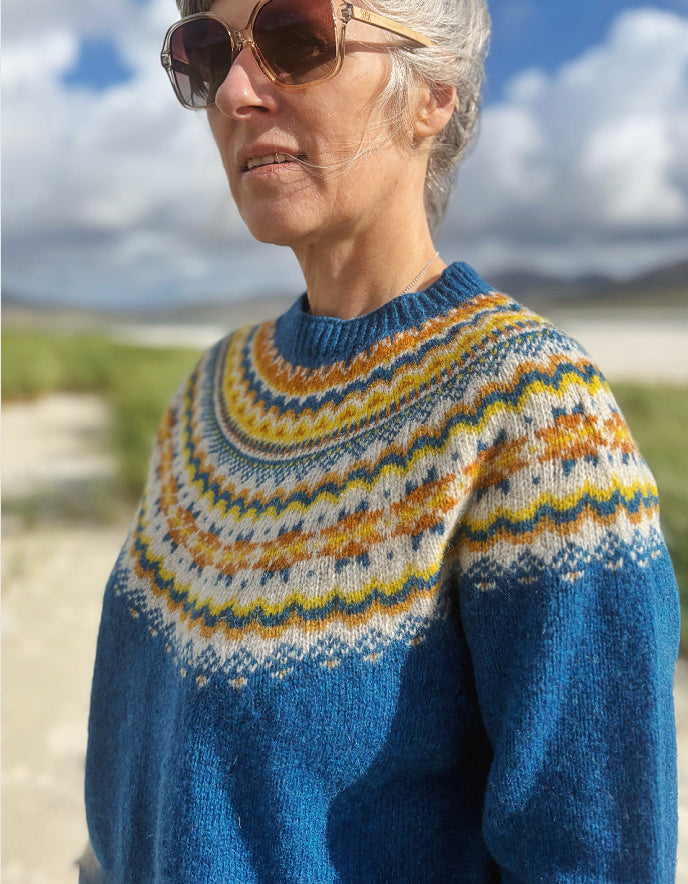 A woman outdoors wearing a blue knit sweater with a colorful Fair Isle yoke pattern in cream, yellow, and orange, along with brown sunglasses.