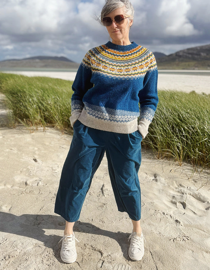 A woman stands on a sandy beach wearing a blue knit sweater with a multicolor Fair Isle yoke, blue cropped pants, and white sneakers, with grass and a cloudy sky in the background.