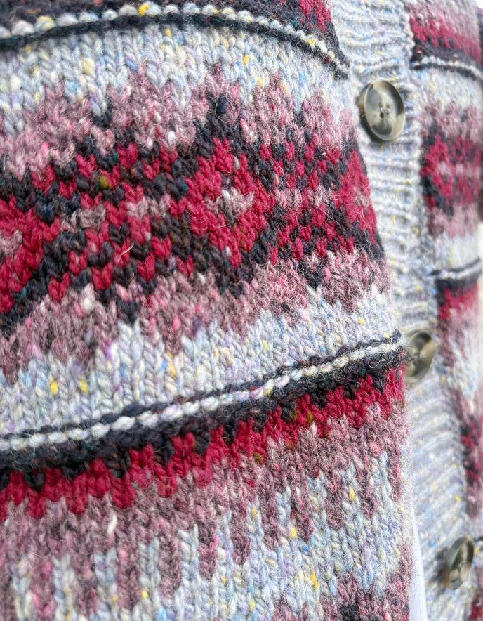 Woman wearing a fair Isle cardigan and plum cord trousers and sunglasses standing by a beach.