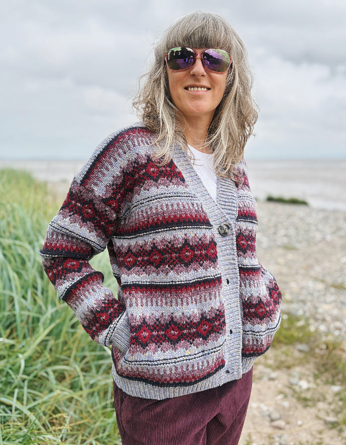Woman wearing a fair Isle cardigan and plum cord trousers and sunglasses standing by a beach.