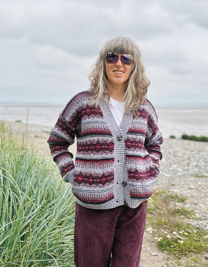 Woman wearing a fair Isle cardigan and plum cord trousers and sunglasses standing by a beach.