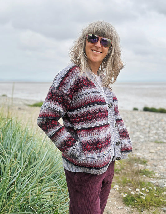 Woman wearing a fair Isle cardigan and plum cord trousers and sunglasses standing by a beach.
