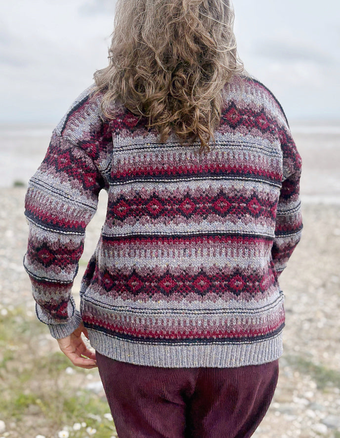 Woman wearing a fair Isle cardigan and plum cord trousers and sunglasses standing by a beach.