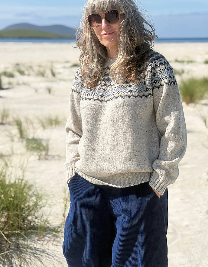 A woman standing outdoors on a sandy beach wearing a cream knit sweater with a dark geometric yoke pattern and dark blue corduroy pants.