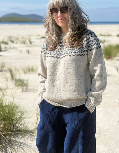 A woman standing outdoors on a sandy beach wearing a cream knit sweater with a dark geometric yoke pattern and dark blue corduroy pants.