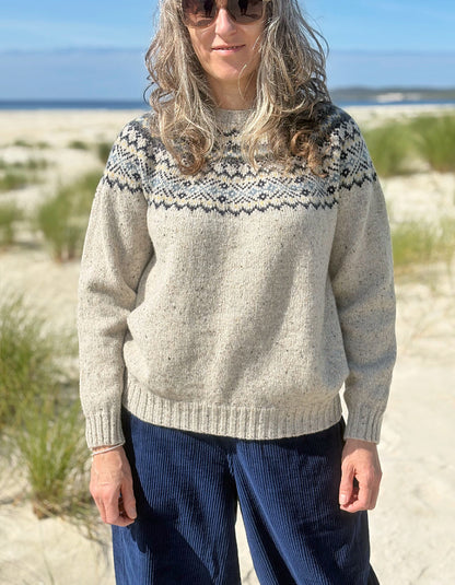 A woman standing on a sandy beach wearing a cream-colored knit sweater with a dark navy and gray geometric Fair Isle yoke pattern, paired with navy blue corduroy pants.