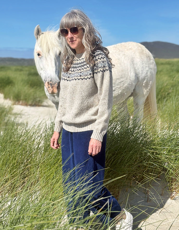 A woman wearing a beige knit sweater with a dark navy geometric yoke pattern and navy wide-leg pants stands outdoors on a sandy path with tall green grass, next to a white horse under a blue sky.