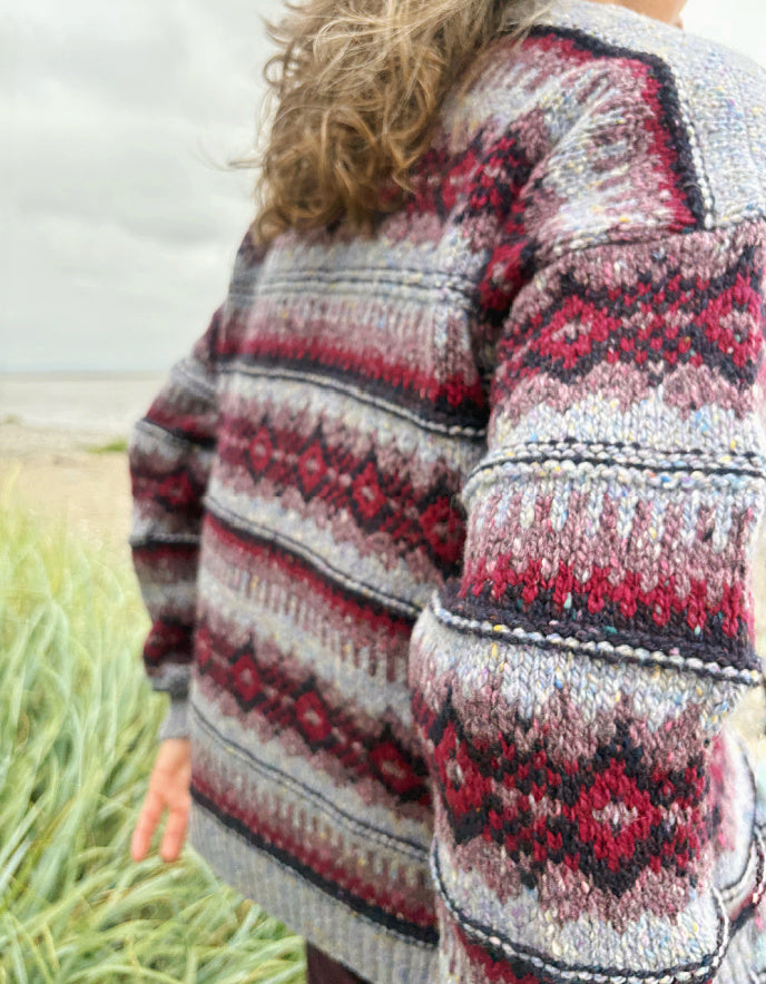 Woman wearing a fair Isle cardigan and plum cord trousers and sunglasses standing by a beach.