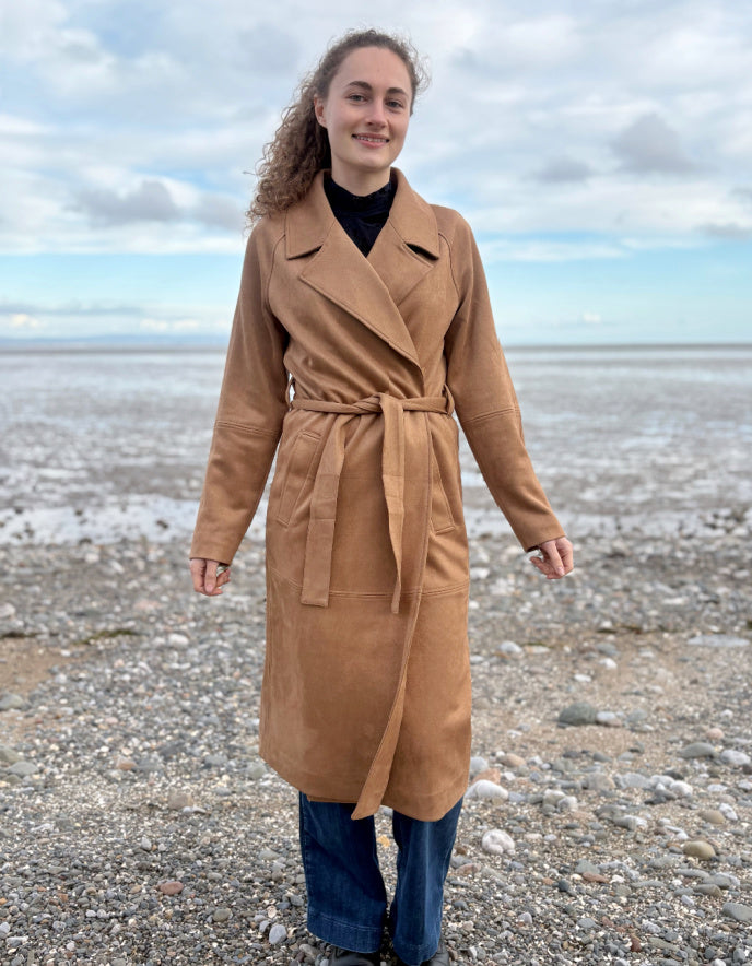 Person wearing a camel faux suede trench coat standing on a beach with a cloudy sky.